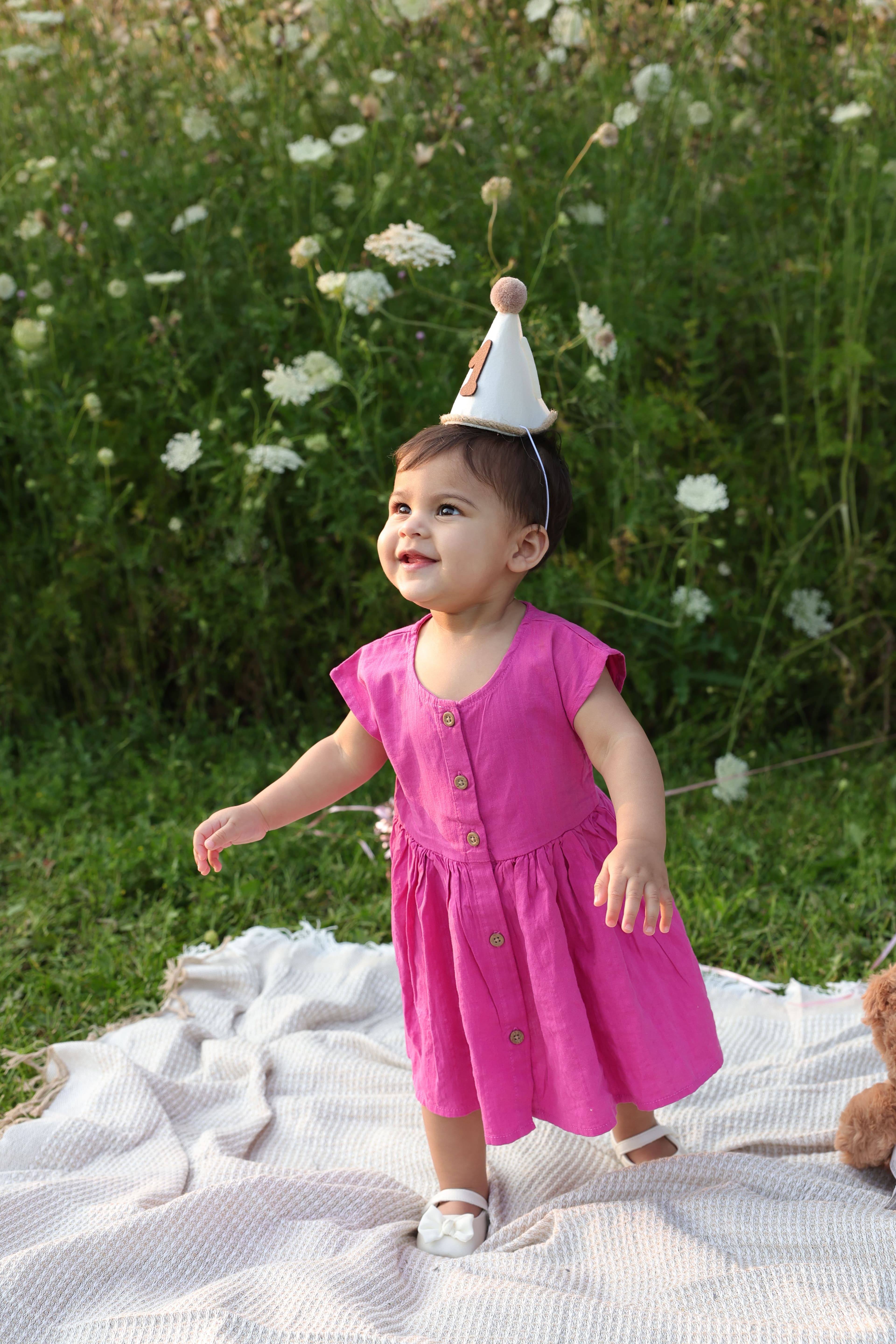 One-year-old smiling in a pink dress and party hat in a wildflower meadow